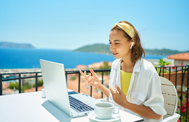 Young mixed race female specialist in wireless headset holding video call on laptop, remote work conference. Millennial woman working during vacation on sea resort