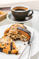 A close-up of a hazelnut and chocolate croissant cut in half on a white plate, a cup of black coffee, a cutlery set, on a glass table on a cafe terrace