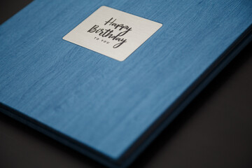 Photobook, photoalbum in blue leather cover on black table background with metallic shield and inscription Happy Birthday to you. Copy space. Soft focused shot, close up.