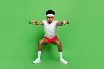 Full length photo of strong serious dark skin guy dressed sport outfit holding dumb bells squatting isolated green color background