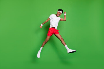 Fototapeta na wymiar Full length photo of impressed excited dark skin man wear white t-shirt rising fists jumping high isolated green color background