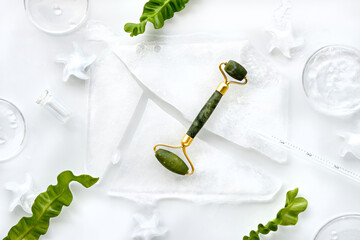 Self made moisturizer and green jade face roller with pieces of ice. Exotic fern leaves and water drops on off white background. Monochromatic white green look. Facial massage, handmade cosmetics. © tilialucida
