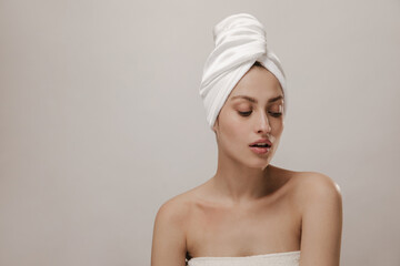 Portrait of handsome girl with white towel wrapped hair and pure cleaned skin, looking down against light background. Wellness and healthcare concept