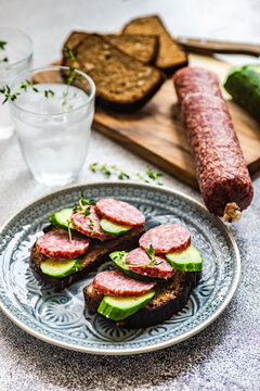 Slices of salami, cucumber and rye bread on a  chopping board with two glasses of water
