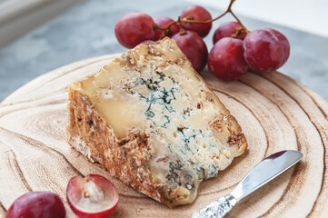 large piece of stilton cheese on a wooden board. Delicatessen cheese with blue mold stilton with grapes and a knife for cutting cheese