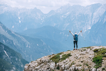 Obraz premium Woman trekker with backpack rose arms up with trekking poles enjoying picturesque Dolomite Alps view near Tre Cime di Lavaredo formation in South Tyrol, Italy. Active people and mountain concept.