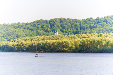 Fototapeta na wymiar A yacht is sailing along the river on a sunny day, a church is on the opposite bank