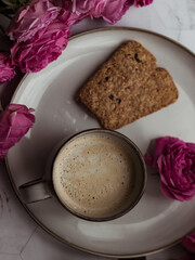 Coffee with milk in a blue ceramic mug, pink roses next to it. Gentle postcard of morning romantic breakfast with coffee.