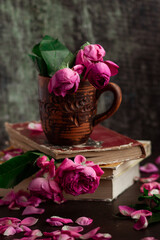 Fascinating pink roses in a brown ceramic mug on a stack of old books; flower petals are scattered nearby. Vintage calm postcard