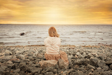 A young woman sits on the rocks by the sea and looks at the sunset