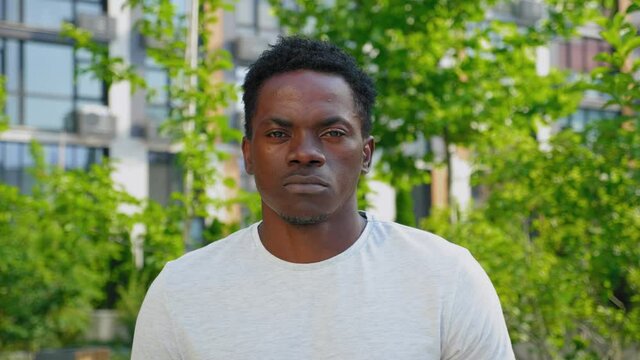Close up African man looks camera on background modern building and trees in summer. Portrait afro-american male wears gray t-shirt stands in park