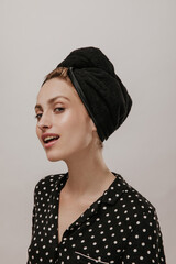 Excited young woman in black polka dot shirt and silk towel on head, looking into camera against light background. Skincare and cleansing concept
