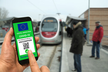 A man at a tram stop holds a smartphone with the European Union digital green pass for Covid-19 in his hand. Safe travel concept during the Coronavirus and the Green pass