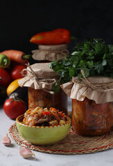 Salad with eggplant, carrot, peppers, garlic, tomatoes in bowl and glass jars on dark background. Vertical format.