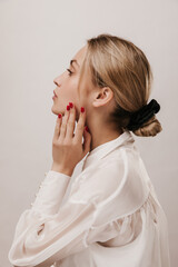 Profile portrait of gorgeous young blonde with white blouse, black silk scrunchie and red manicure seriously looking aside and touching neck. Girl posing isolated over light background