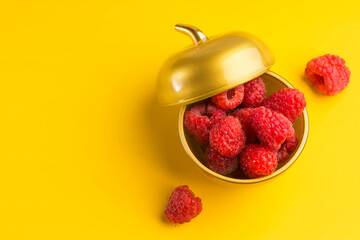 A bowl of gold color with red fresh raspberries on a yellow background. Sweet natural summer harvest. Copy space