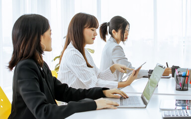 Asian smart young beautiful businesswomen wearing formal suits, working together with intention, using laptop, tablet and notebook to write minute meeting while sitting in modern indoor office.