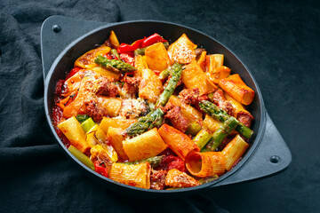 Traditional roasted Italian pasta mezzi paccheri rigati con salsiccia sausage with green asparagus tips in tomato sauce served as close-up in cast iron skillet pan in matte flat cinematic modern look