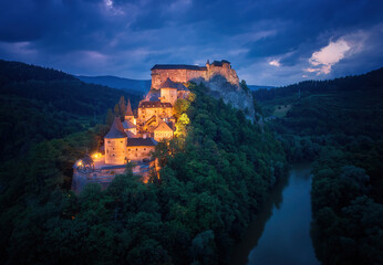 Obraz premium Aerial, dramatic view of Orava Castle situated on a high rock above Orava river, illuminated by orange lamps against dark blue background. Orava, Slovakia.