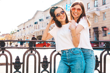 Two young beautiful smiling hipster female in trendy summer white t-shirt clothes and jeans.Sexy carefree women posing on the street background. Positive models having fun, chatting and communicating