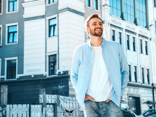 Portrait of handsome smiling stylish hipster lambersexual model.Modern man dressed in blue shirt. Fashion male posing near skyscraper on the street background. Outdoors at sunset