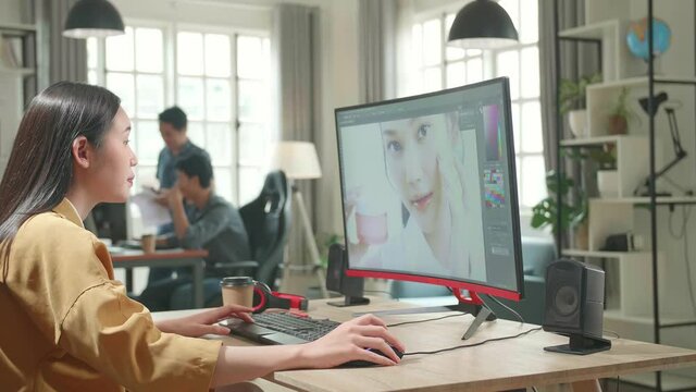 Asian Female Digital Editor Drinking Coffee While Works In Photo Editing Software On Her Personal Computer With Big Display In Office
