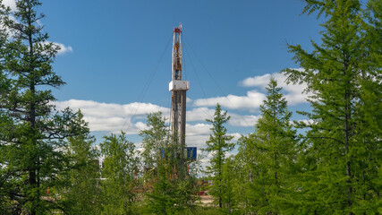Obraz premium Landscape with a drilling rig in an oil and gas field. Infrastructure, communications and drilling equipment are visible. Around the rig there is summer northern taiga with green trees and blue sky