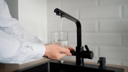 Man fills an empty glass with drinking water in the kitchen. Water flows from the tap into a transparent container. A sink, a tap, male hands and a glass are visible in the shot