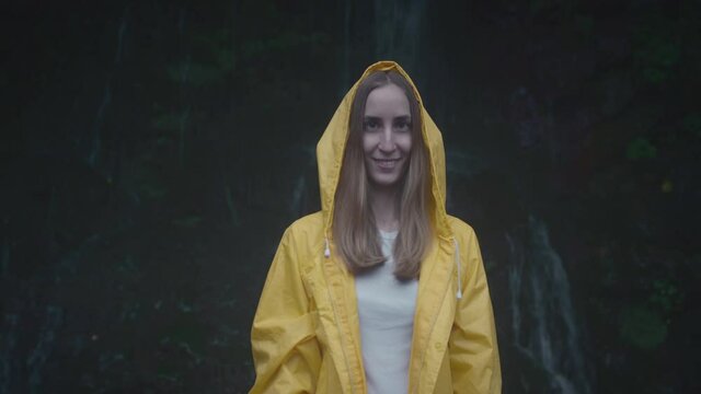 Portrait of Caucasian Girl traveler in a yellow raincoat near a waterfall in the forest. Woman tourist enjoying waterfall. Traveling in mountains, adventure in trip. Lifestyle concept, in slow motion