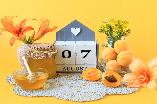 Calendar for August 7 : the name of the month of August in English, the numbers 0 and 7, flowers in vases, a jar of jam, apricots on a gray napkin, yellow background, side view