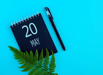 May 20th. Day 20 of month, Calendar date. Black notepad sheet, pen, fern twig, on a blue background. Spring month, day of the year concept