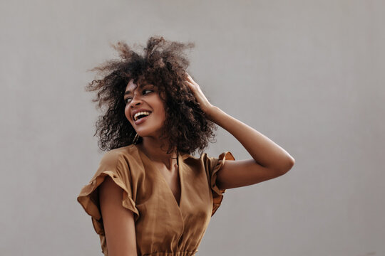 Excited lady in brown blouse touches hair and looks back. Attractive dark-skinned woman smiles outside.