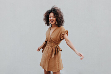 Excited dark-skinned lady in beige dress smiles and turns on grey background. Curly brunette woman...