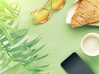 Flat lay of paper cup of coffee, croissant, phone, sun glasses and palm leaves on green background, bleached colors. The rays of sun pass through leaves of palm tree. Breakfast concept. Copy space.
