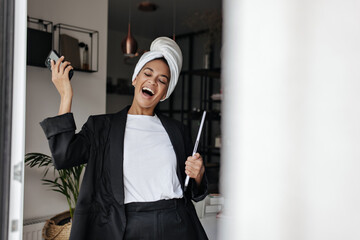 Excited emotional dark-skinned woman in white t-shirt and black suit rejoices, holds laptop and smartphone.