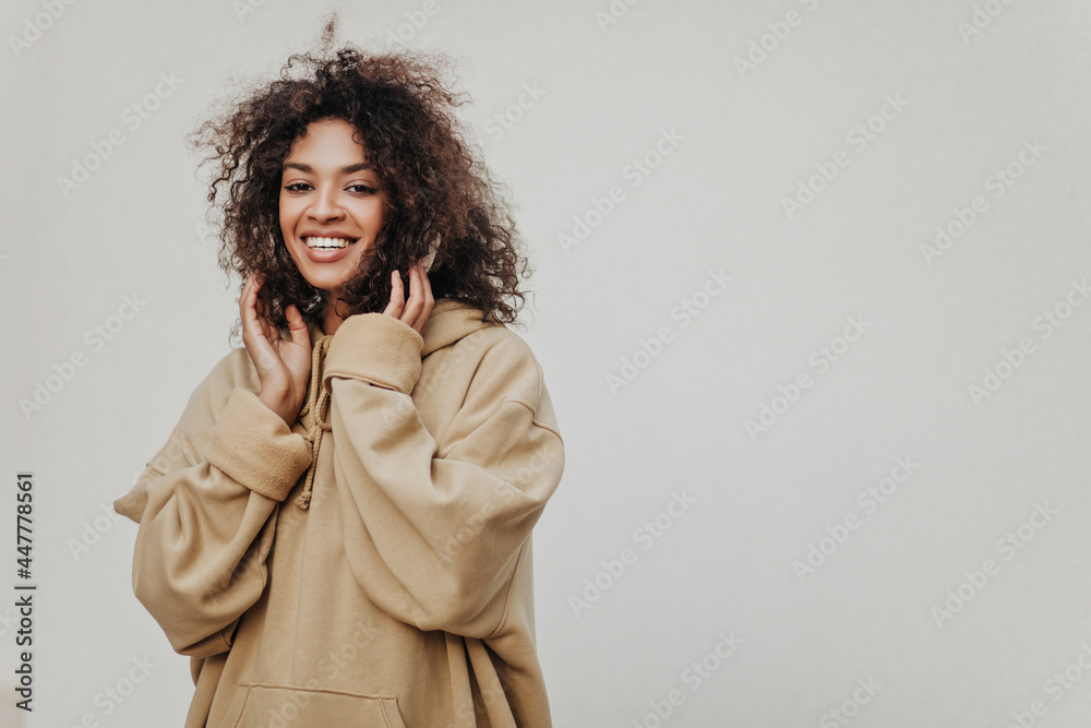 Poster Curly brunette African woman listens to music in headphones on grey background. Dark-skinned girl in hoodie smiles.