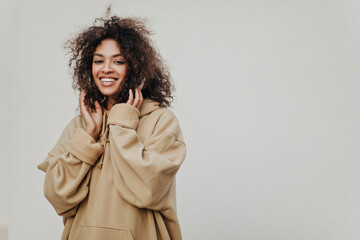 Curly brunette African woman listens to music in headphones on grey background. Dark-skinned girl in hoodie smiles.