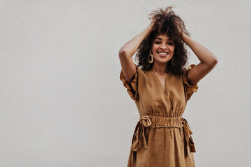 Dark-skinned woman in brown dress smiles sincerely and ruffles hair outside. Curly lady looks into...