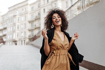 Pretty brunette woman laughs and walks outside. African dark-skinned lady in brown dress and coat moves and smiles outdoors. - obrazy, fototapety, plakaty
