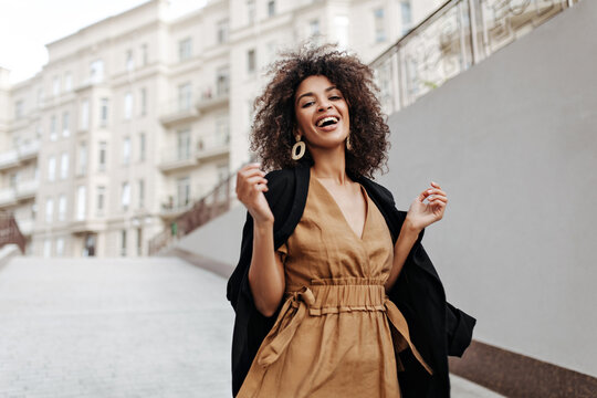 Pretty brunette woman laughs and walks outside. African dark-skinned lady in brown dress and coat moves and smiles outdoors.