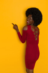 Young Black Woman In Elegant Red Mini Dress Is Pointing At Yellow Wall