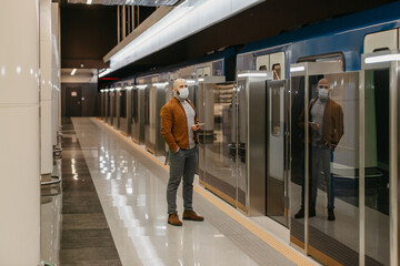 A man in a face mask is holding a smartphone while waiting for a subway train.