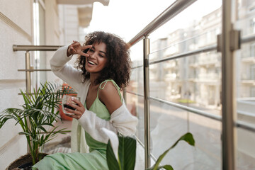 Brunette woman shows peace sign and holds coffee glass. Beautiful lady in silk dress and white cardigan sits on balcony.