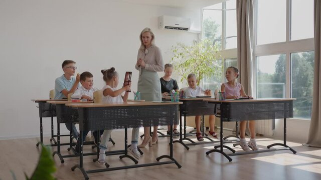 In Elementary School Class: Enthusiastic Teacher walks between Rows of Bright Diverse Children, Explains Lesson. Group of Smart Multiethnic Kids Learning New Stuff. Side View Tracking Shot