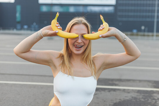 Frivolously and mischievous young woman with braces on teeth holding bananas on her face has fun outdoors