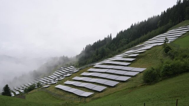 Solar park, photovoltaic power station located on a mountain slope in the Alps. Foggy, rainy day, green pasture grass and mountains create a fresh, ecological image.