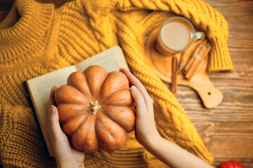 Ripe pumpkin in women's hands over knitted sweater. Hot cocoa with cinnamon sticks on wooden tray....