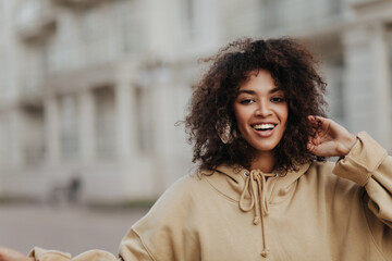 Dark-skinned brunette woman listens to music in headphones outdoors. Curly lady in beige hoodie smiles outside.