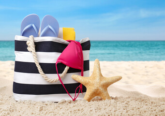 Stylish bag with different accessories on sandy beach near ocean