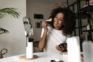 Joyful young brunette woman in white t-shirt smiles, powders face, holds palette abc makeup brush, looks into mirror.